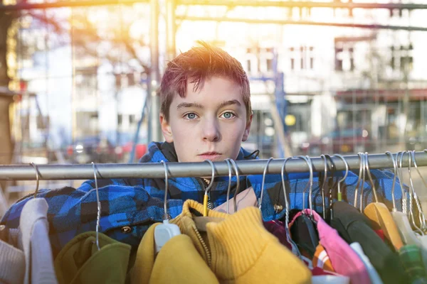 Portrait of a teenage boy behind a clothes rail on a flea market