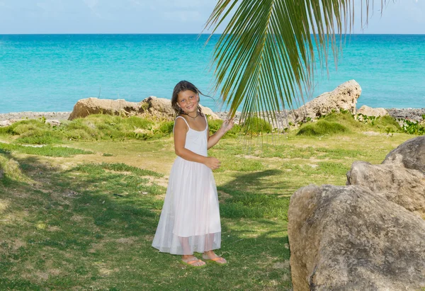 Smiling joyful little girl standing in gorgeous tropical garden against azure tranquil ocean and blue sky background