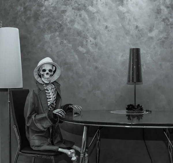 Happy skeleton sitting behind the table in dark dramatic room environment, wearing leather jacket and white hat and waiting for Halloween party
