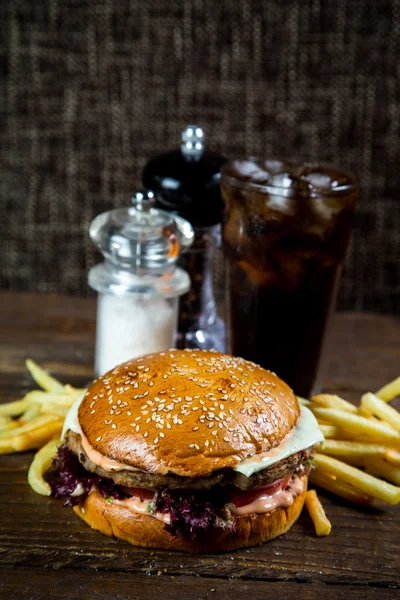 Big and tasty hamburger on desk with cola and fried potatoes on dark background