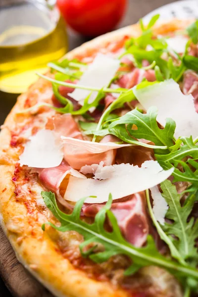 Pizza with prosciutto (parma ham), arugula (salad rocket) and parmesan on dark wooden background close up. Italian cuisine.