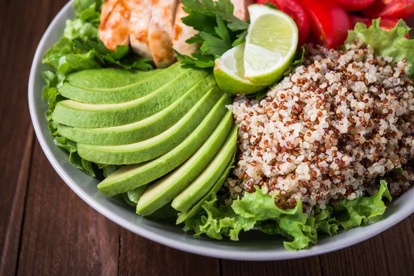 Healthy salad bowl with quinoa, tomatoes, chicken, avocado, lime and mixed greens (lettuce, parsley) on wooden background close up.