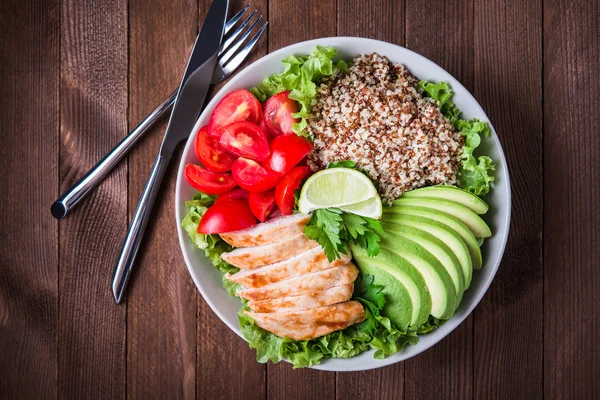Healthy salad bowl with quinoa, tomatoes, chicken, avocado, lime and mixed greens (lettuce, parsley) on wooden background top view.