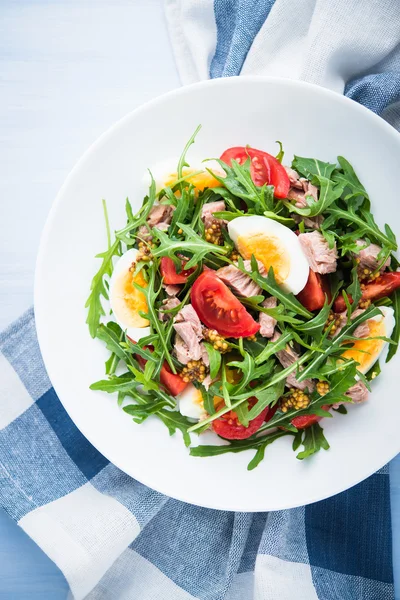 Fresh salad with tuna, tomatoes, eggs, arugula and mustard on blue wooden background top view.