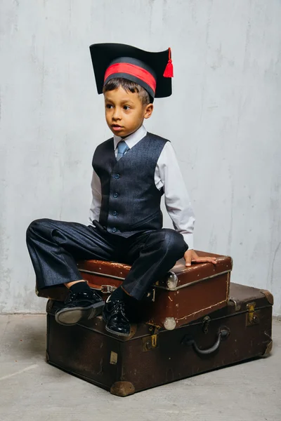 Small black boy in  graduation hat sits on the old suitcases