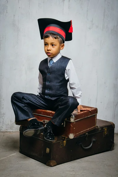 Small black boy in  graduation hat sits on the old suitcases
