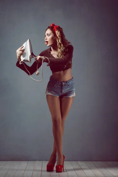 Pin up Young beautiful sexy girl with make-up blonde with a red band in denim shorts housewife with iron in red shoes slender legs