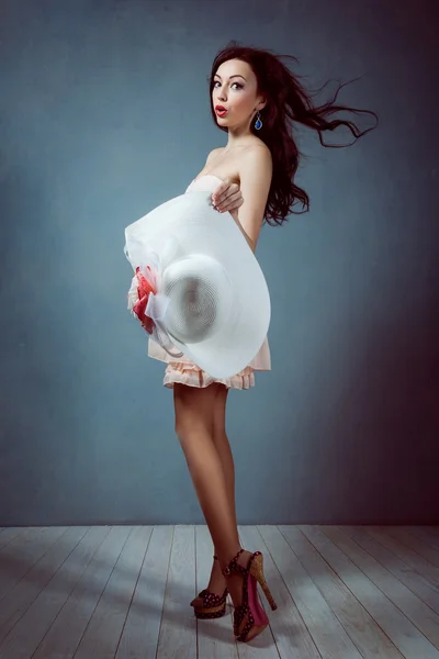 Young beautiful sexy brunette girl with long flowing hair holding a hat with a make-up with a beautiful figure in a pink dress with long legs in shoes and heels pin up