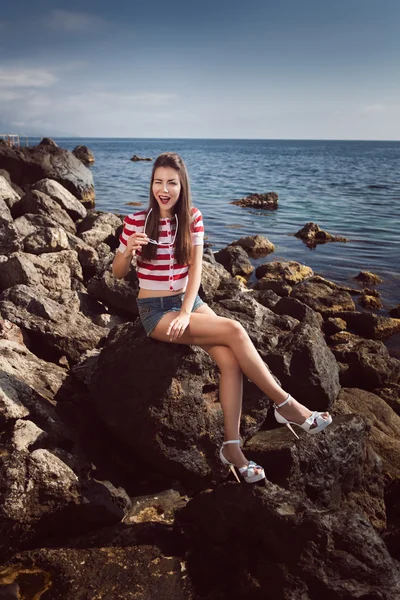 Pin up young beautiful girl on the sea in short jeans and striped T-shirt with long hair rocks glasses gulls water clouds, wave, dream figure with glasses emotions, smile with teeth, open mouth