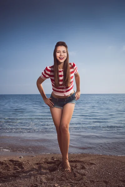 Pin up young beautiful girl on the sea in short jeans and striped T-shirt with long hair rocks glasses gulls water clouds, wave, dream figure with glasses emotions, smile with teeth, open mouth