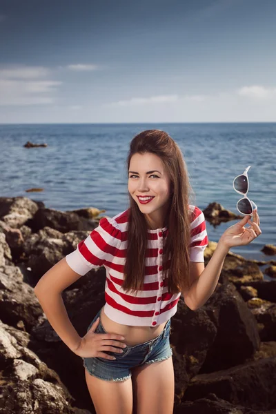 Pin up young beautiful girl on the sea in short jeans and striped T-shirt with long hair rocks glasses gulls water clouds, wave, dream figure with glasses emotions, smile with teeth, open mouth