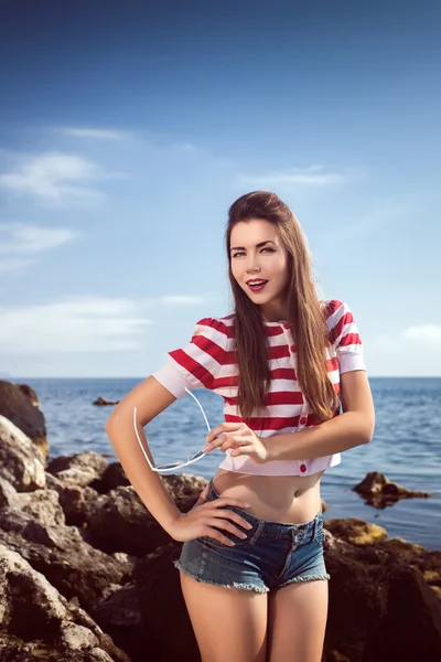 Pin up young beautiful girl on the sea in short jeans and striped T-shirt with long hair rocks glasses gulls water clouds, wave, dream figure with glasses emotions, smile with teeth, open mouth