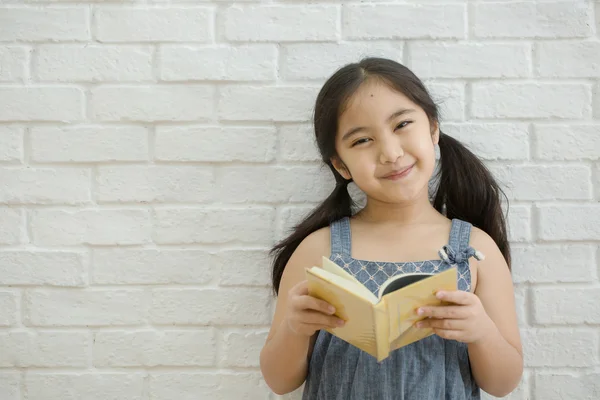 Happy Asian girl reading story book on white brick wall