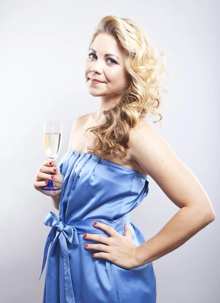 Beautiful curly-haired blond with a glass of champagne. A young woman in blue evening dress. Smiling girl on a gray background