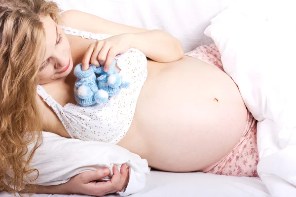 Beautiful pregnant blonde woman with booties in her hand.young pregnant woman luxuriates in a white bed.Woman Bed Portrait.Beautiful pregnant curly blonde