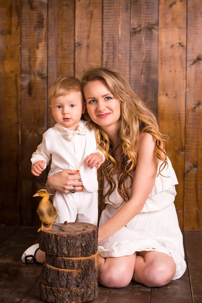 Baby blond boy in a white suit white socks standing with his mother on a wooden background. next to a small duckling