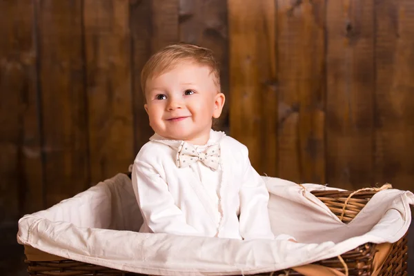 Baby blond boy in a white suit white socks sitting in basket on a wooden background. next to a small duckling