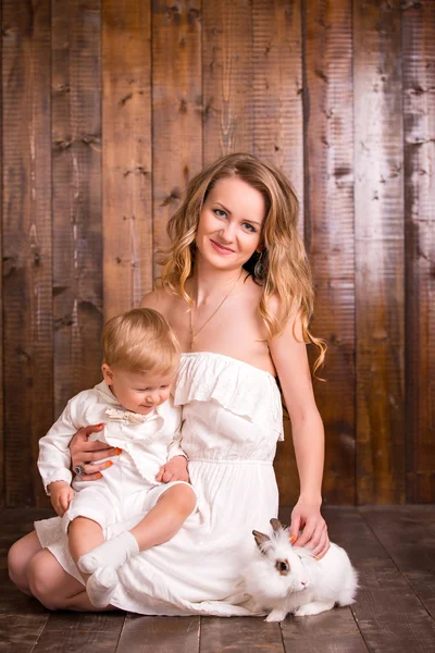 Baby blond boy in a white suit white socks with his mother on a wooden background. next to a small duckling