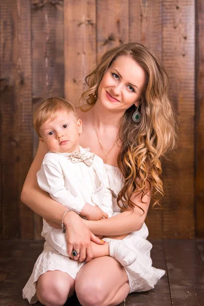 Baby blond boy in a white suit white socks with his mother on a wooden background. next to a small duckling