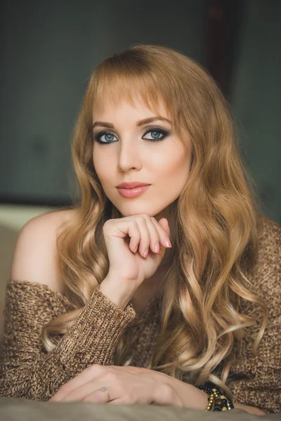 Young beautiful girl blonde woman in a long pullover, beautiful wavy hair, long legs, a wooden background, bed