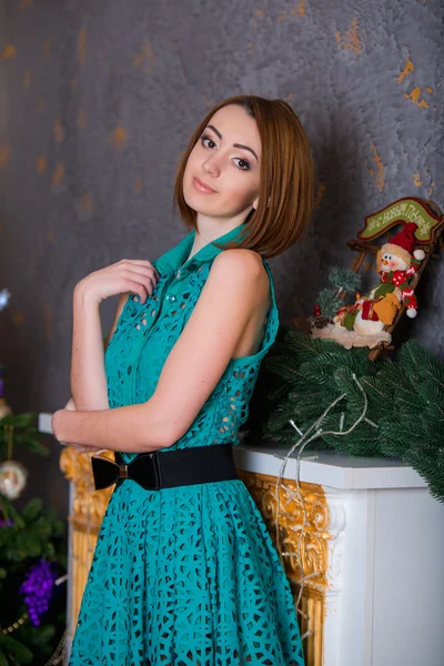 Beautiful young girl sitting on the floor on a background of green trees, she is dressed in a green dress. She smiles, she has red hair, big eyes, she stands by the fireplace