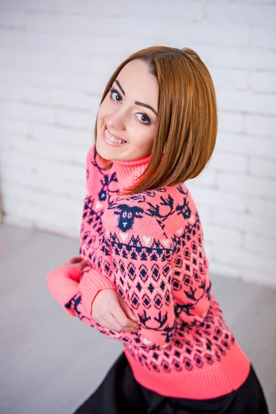 Beautiful young girl sitting on the floor against a white brick wall, she is dressed in a pink sweater, black skirt, socks. Girl smiles, she has red hair, big eyes