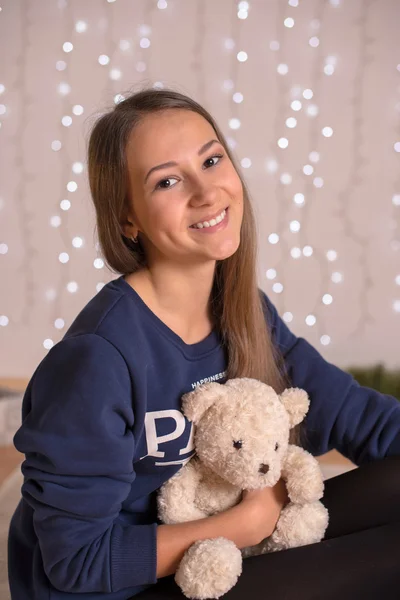 Beautiful young girl in a sweatshirt and tights near the Christmas tree, Christmas lights in the background, she smiles, happy, looking directly,white socks, warm socks, a white teddy bear, hugging a pillow,