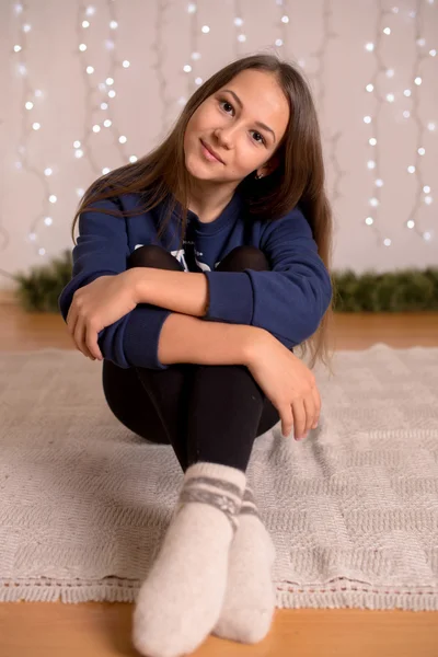 Beautiful young girl in a sweatshirt and tights near the Christmas tree, Christmas lights in the background, she smiles, happy, looking directly,white socks, warm socks, a white teddy bear, hugging a pillow,