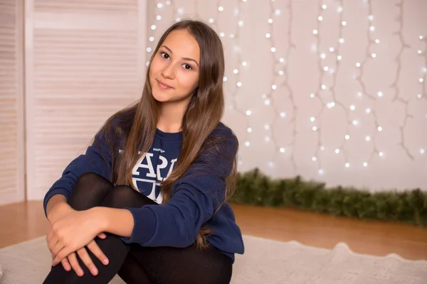 Beautiful young girl in a sweatshirt and tights near the Christmas tree, Christmas lights in the background, she smiles, happy, looking directly,white socks, warm socks, a white teddy bear, hugging a pillow,