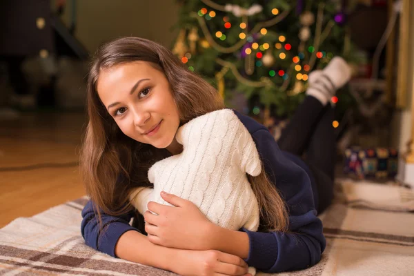 Beautiful young girl in a sweatshirt and tights near the Christmas tree, Christmas lights in the background, she smiles, happy, looking directly,white socks, warm socks, a white teddy bear, hugging a pillow,