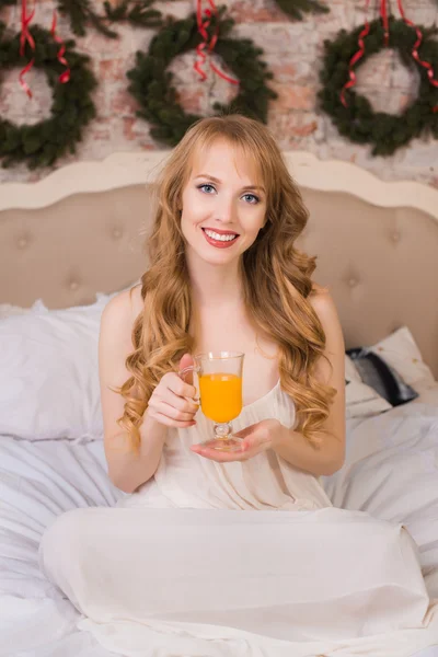 Beautiful young blonde woman with long hair in a white nightie simple dress sitting on a bed and holding a transparent glass with orange juice, morning, christmas, new year, holiday, happiness