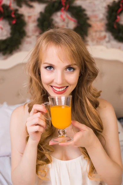 Beautiful young blonde woman with long hair in a white nightie simple dress sitting on a bed and holding a transparent glass with orange juice, morning, christmas, new year, holiday, happiness