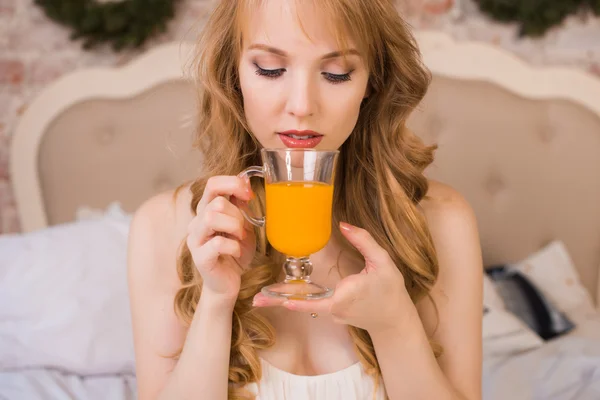 Beautiful young blonde woman with long hair in a white nightie simple dress sitting on a bed and holding a transparent glass with orange juice, morning, christmas, new year, holiday, happiness