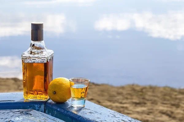 Bottle tequila and tumbler with lemon on shore of lake