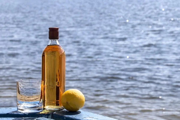 Bottle whiskey and tumbler with lemon on shore of lake