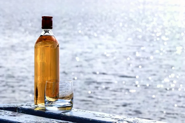 Bottle whiskey and tumbler on shore of lake