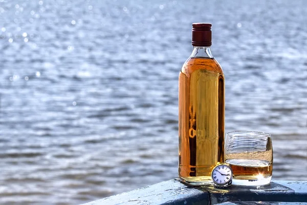 Bottle of whiskey and tumbler with a watch on shore of lake