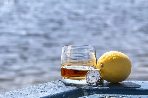 Tumbler whiskey and lemon with watch on shore of lake