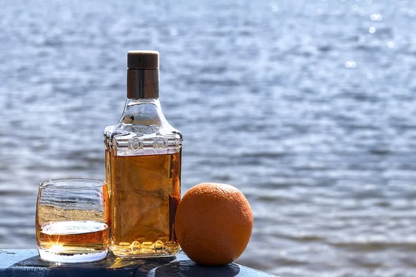 Bottle of tequila and tumbler with Orange on the shore of lake