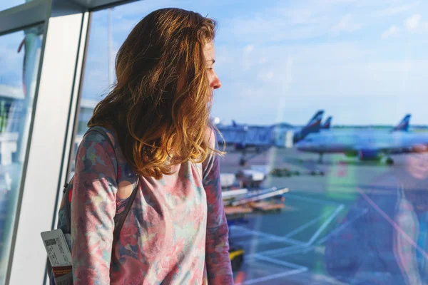 Pretty young girl traveler with a backpack next to the panoramic windows of the passenger terminal  international airport watching the planes on the airfield.