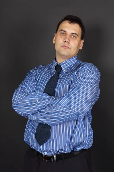 Handsome young cross-armed man in tie and blue shirt stands in b