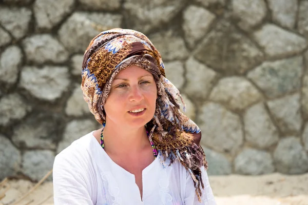 Portrait of middle-aged woman in summer at the beach. Scarf tied around the head as a turban. Against stone wall as a background.