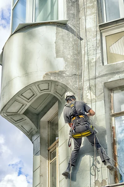 Worker alpinist in a protective mask working with sander for smoothing wall surface of the building