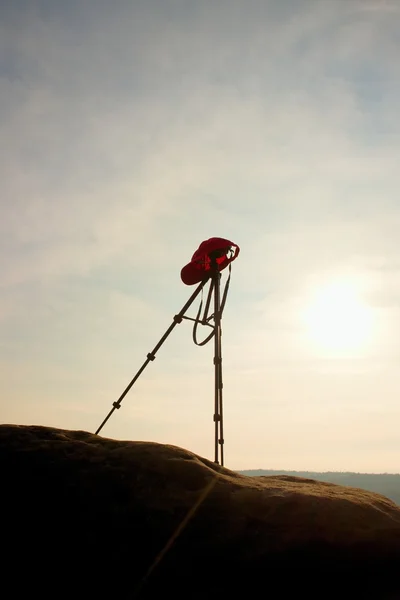 Tripod with red baseball cap on the peak ready for photography. Sharp autumn rocky peaks increased from gold fog.