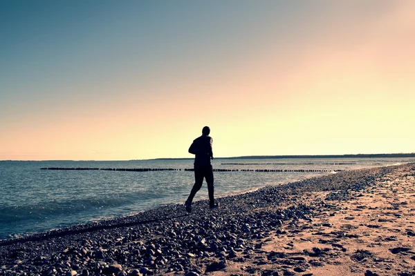 Tall man in dark sportswer running and exercising on stony beach at breakwater.