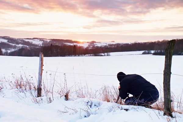 Photographer lay in snow on meadow and takes a picture of fantastic sunset above hill