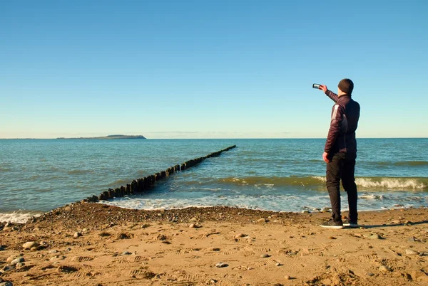 Tall man hold cellphone, take picture of autumn  sea scenery at breakwater.