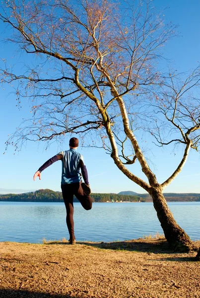 Silhouette of sport active man in running leggins and blue shirt at birch tree on  beach. Calm water, island and sunny day