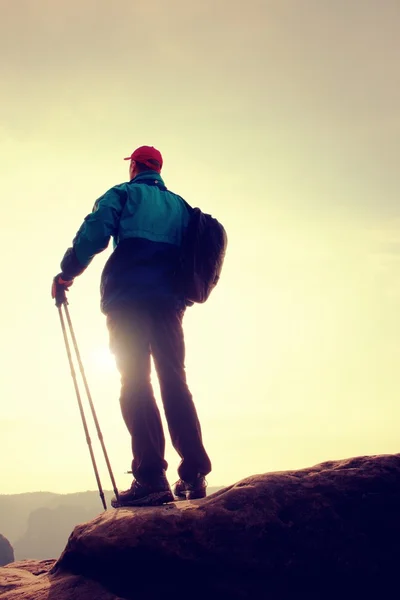 Hiker in windcheater, baseball cap and with trekking poles stand on mountain peak rock.