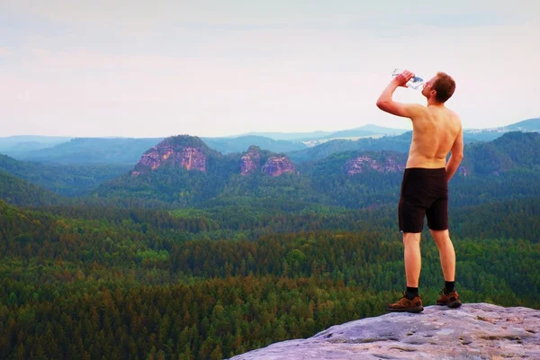 Thirsty hiker in black pants with bottle of water. Sweaty tired tourist on the peak of sandstone rocky park Saxony Switzerland watching into misty landscape.